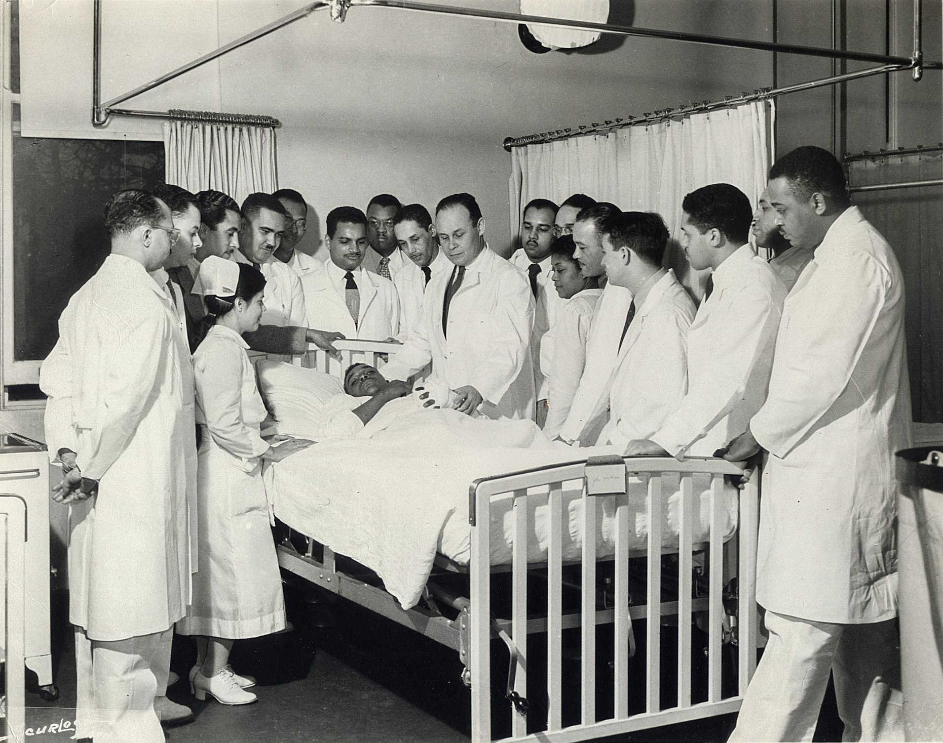 Dr. Charles Drew inspects a patient’s injured hand while dozens of doctors in white lab coats observe. 