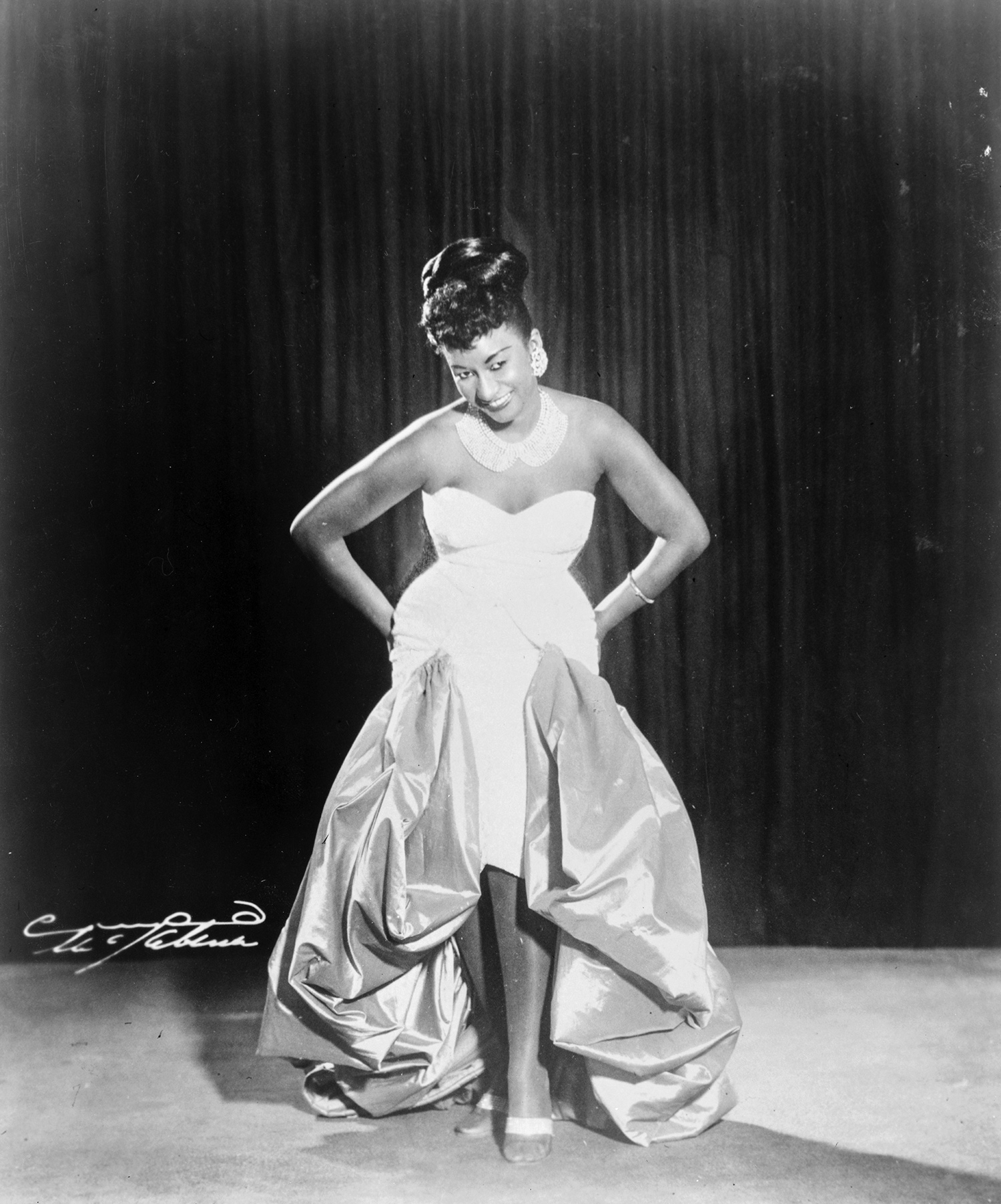 Un retrato fotográfico de cuerpo entero de una mujer con un glamuroso vestido de noche y sonriendo con la mirada baja.