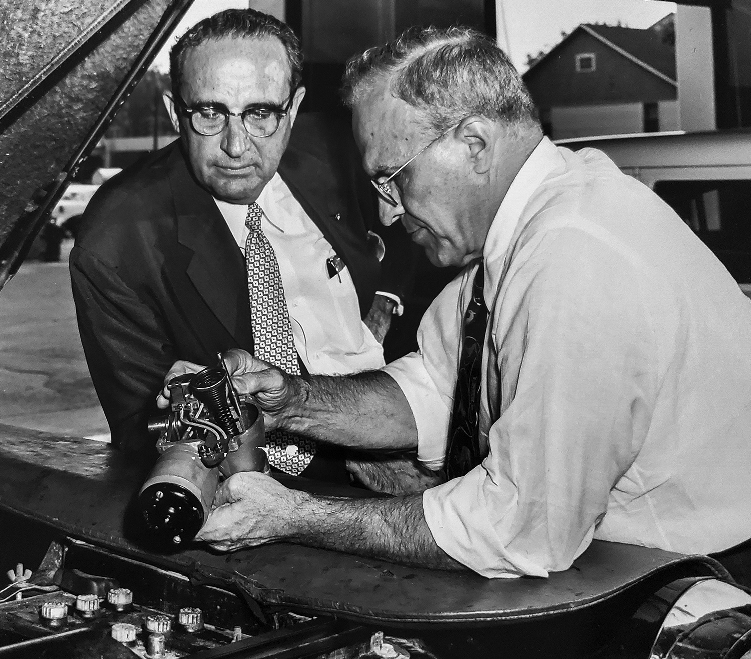 Two men lean on the hood of a car while man in white dress shirt explains device he is holding. 