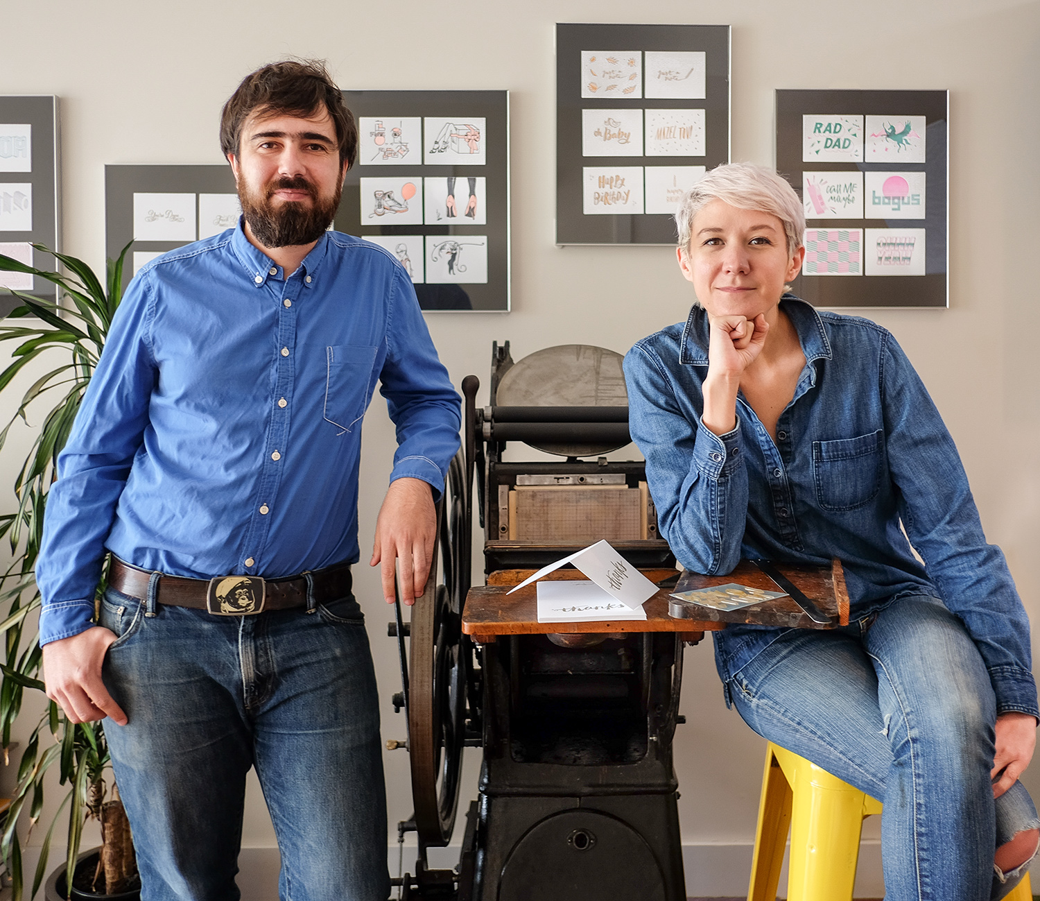 Lex and Santiago pose on either side of a vintage letter press with framed cards in the background. 