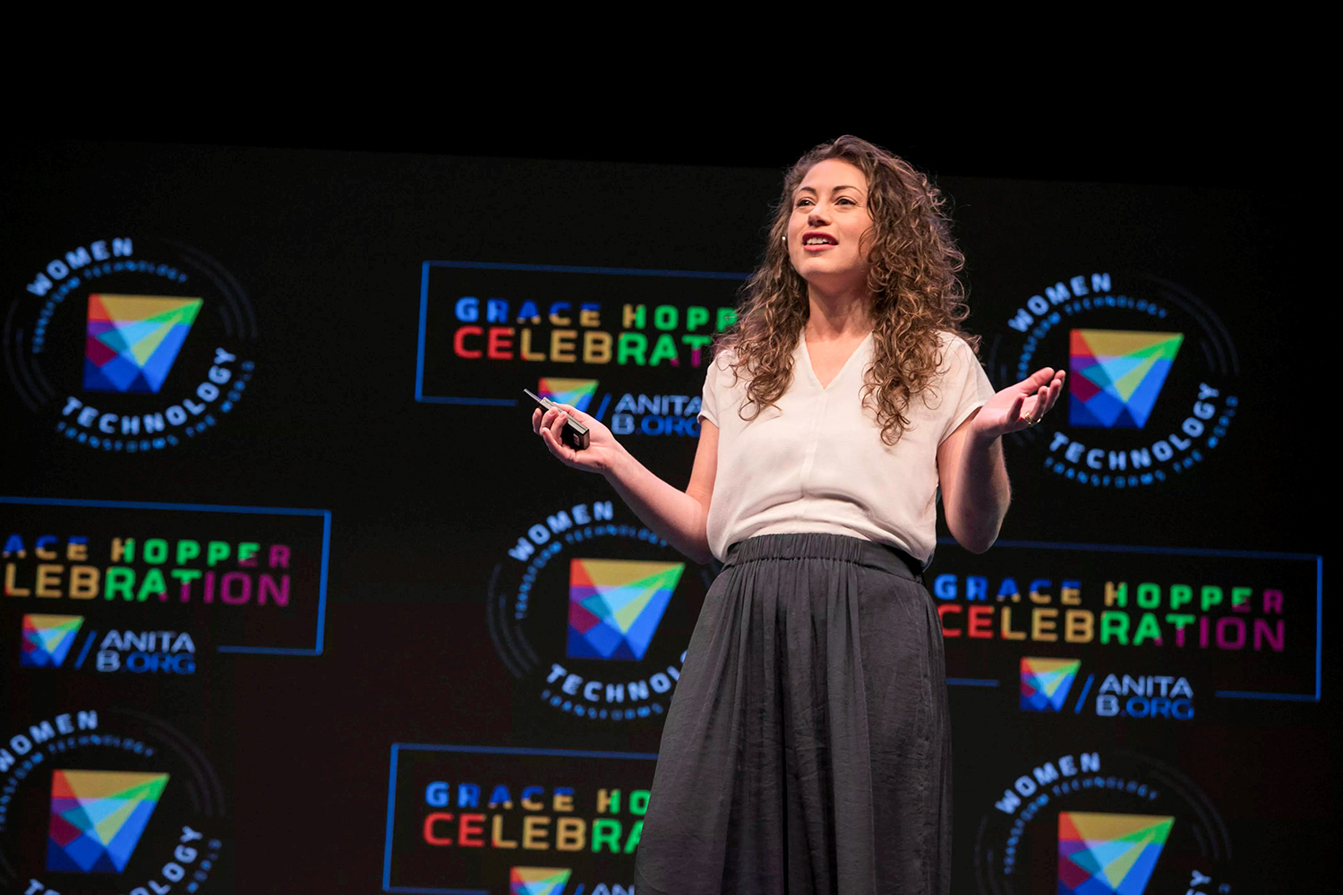 Sterling standing and speaking with a slight smile in front of a black backdrop with colorful logos for the Grace Hopper Celebration of Women in Computing. 