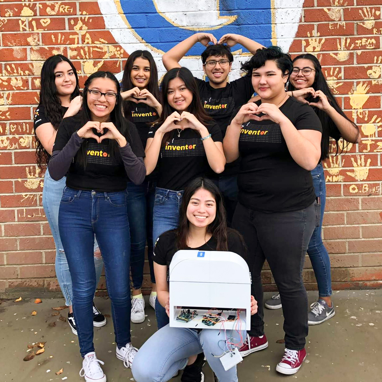 7 diverse students making heart shape sign with hands and one female student in front kneeling and holding an invention and smiling