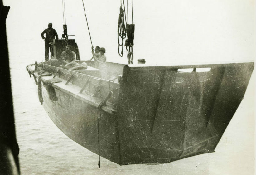 Image: Soldiers standing in a Higgins boat, which is being lowered into the water on ropes from a larger navy ship.