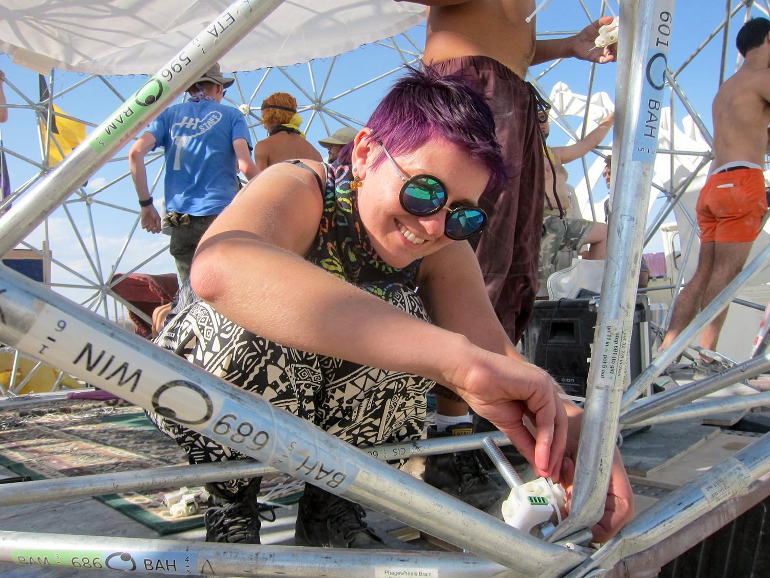  Natalia Bilenko in a pink hard hat and with lab goggles around her neck, holds up and admires a fist-sized clear plastic prototype of Dr. Brainlove. Behind her, workers in hard hats assemble the large metal Dr. Brainlove sculpture. 