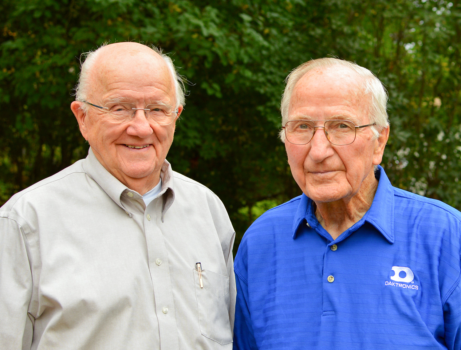 Duane Sander and Al Kurtenbach standing next to each other and smiling in front of dark green pine trees. Sander wears a tan dress shirt without tie and Kurtenbach wears a blue polo shirt with a Daktronics logo. 2023. Photo by David Kadas/USPTO.