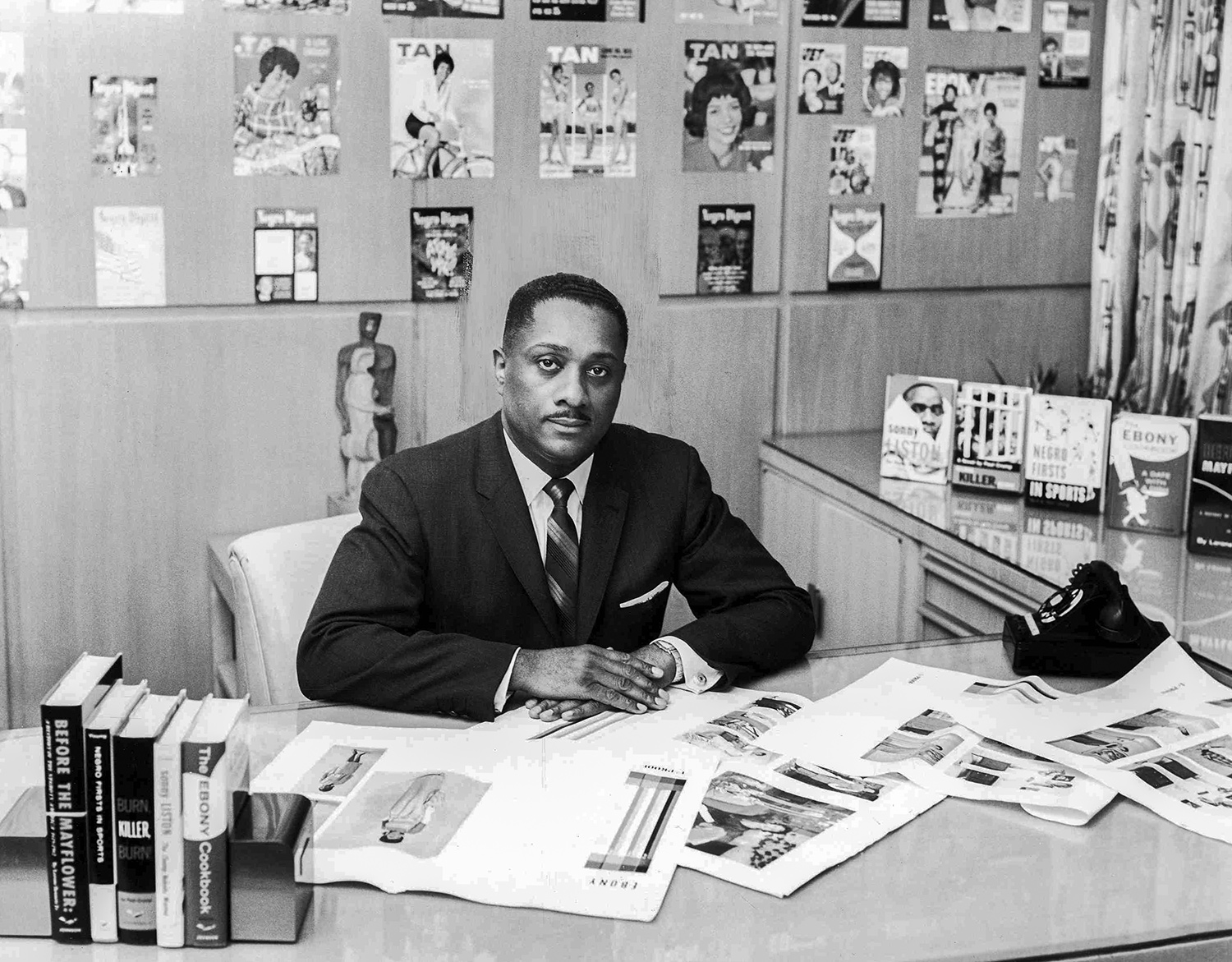 Man wearing a dark suit and tie sitting at his office desk that is covered in open magazines.