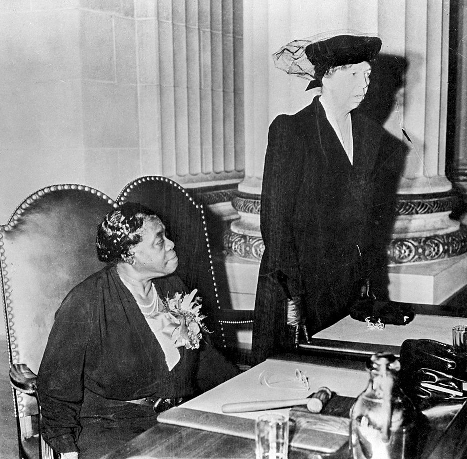 Black and white image of a woman wearing a dark dress sitting at a desk with a woman wearing a black dress suit and hat standing next to the office desk.