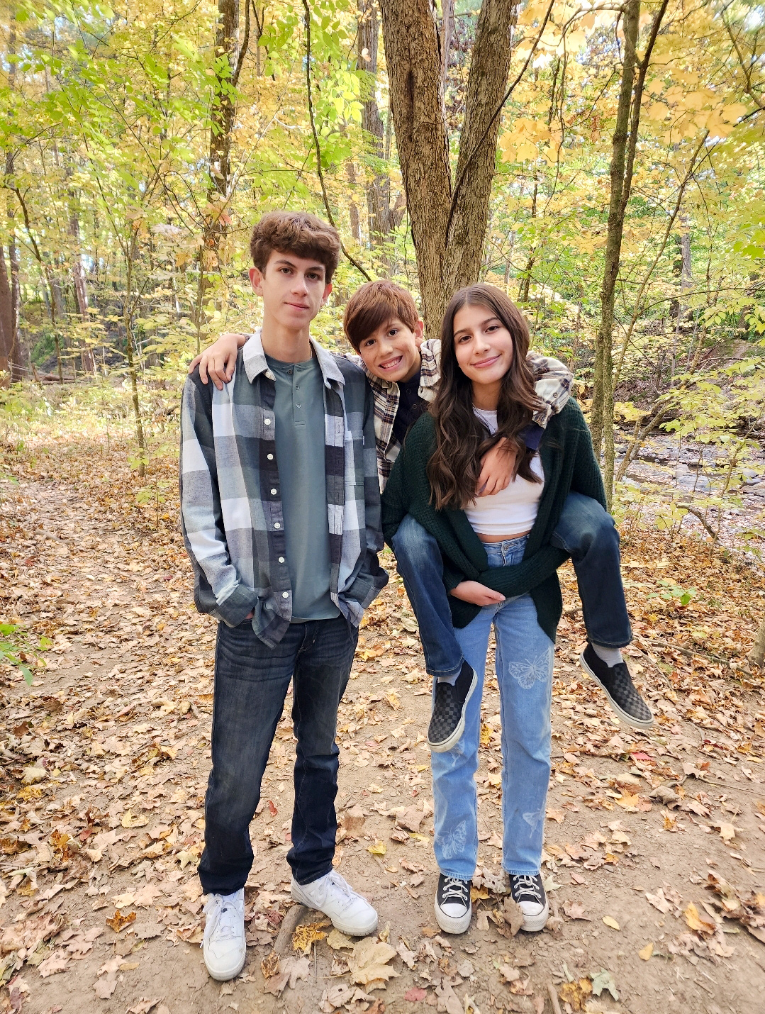 Three school-age children, two boys and one girl) stand together on a nature trail with woods in the background.