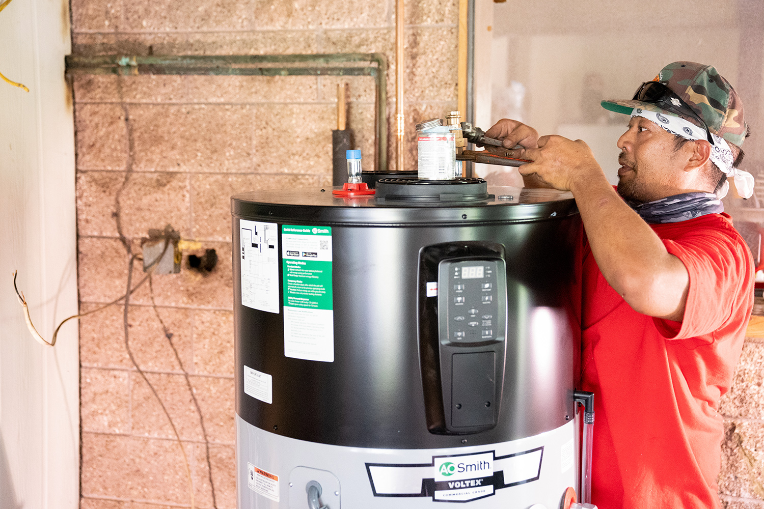 Man in hat and red shirt is using tools to install a new electric water heater