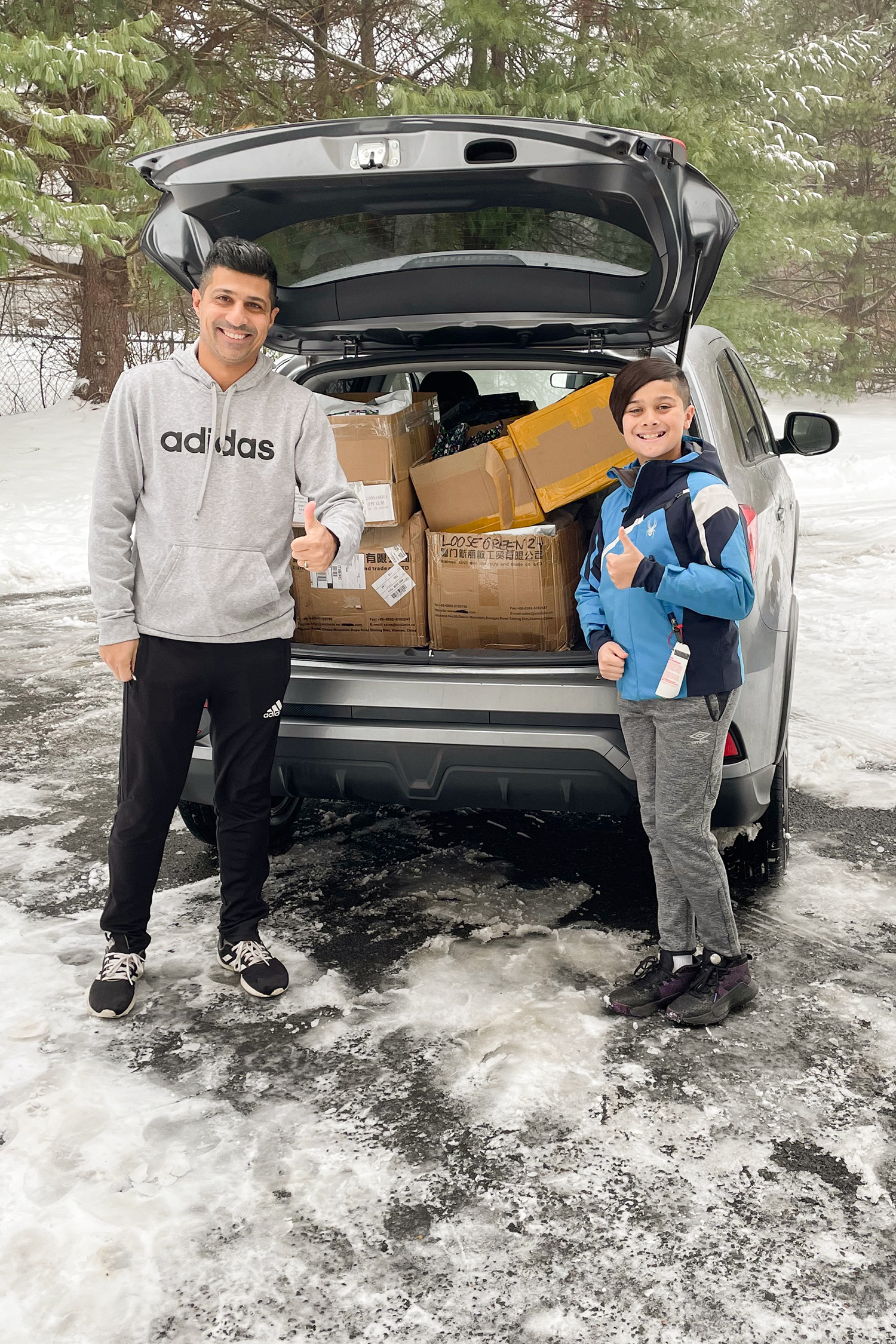 A man and his son standing behind a car filled with boxes, both show a thumbs up sign