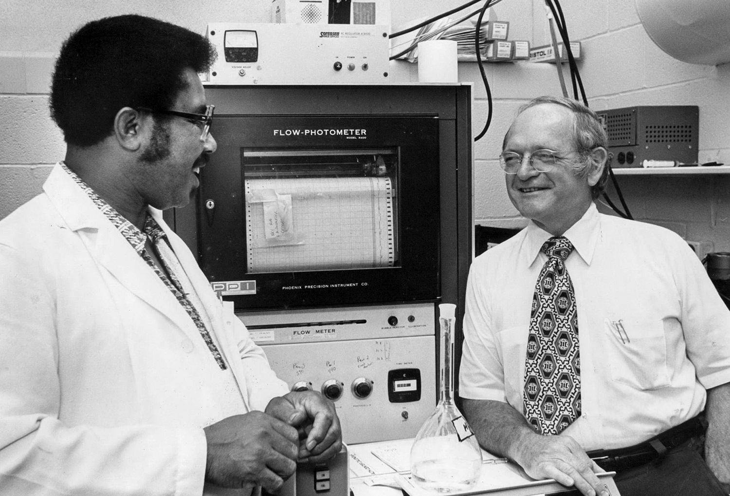 Man with glasses and white lab coat talks with older man 