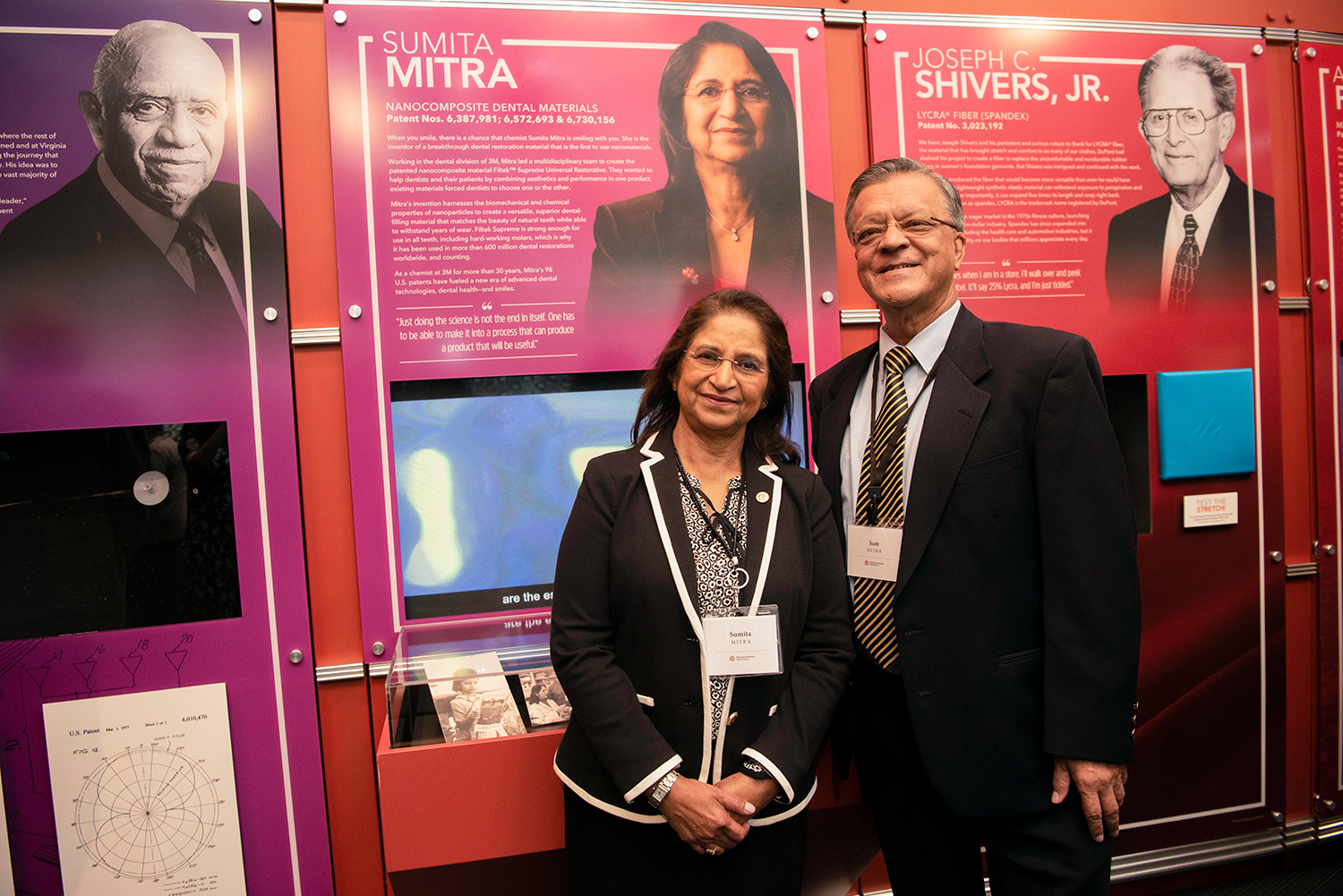 Woman wearing a black pants suit stands next to a man wearing a suit and tie in front of an educational display