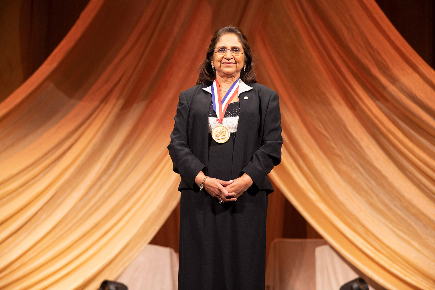 Woman wearing long black dress and suitcoat stands and displays medal from National Inventors Hall of Fame