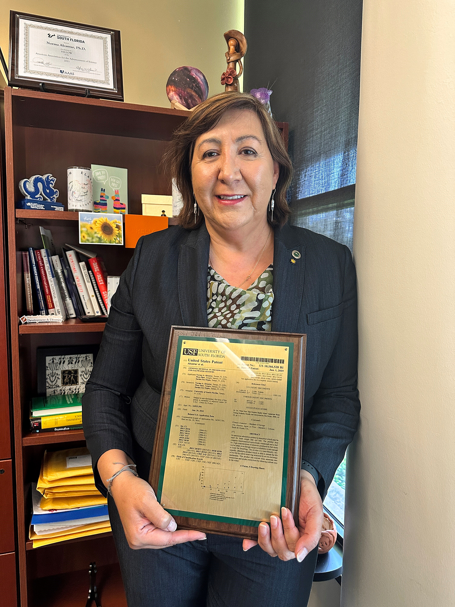 Una profesora universitaria vestida con un traje negro sostiene una placa que representa una patente que le fue concedida.