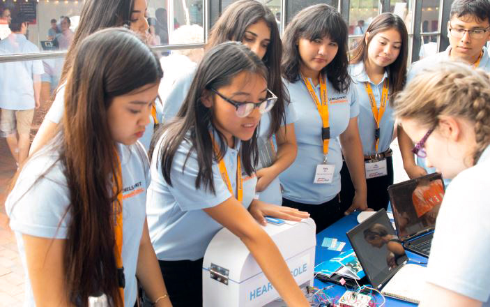 Varios estudiantes de secundaria con camisas azul celeste interactúan con una persona en una mesa de exhibición. Frente a ellos hay un dispositivo blanco, dispositivos electrónicos y varias computadoras portátiles.