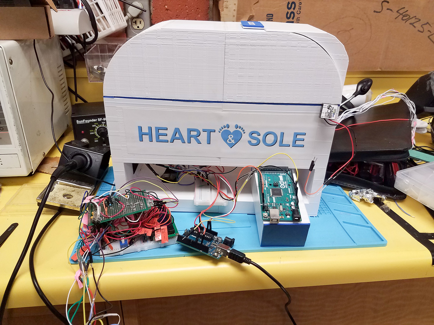 Una caja blanca, similar a una CPU, se muestra en una mesa de trabajo de un laboratorio con elementos electrónicos expuestos. Está etiquetada “Heart and Sole” con letras azules.