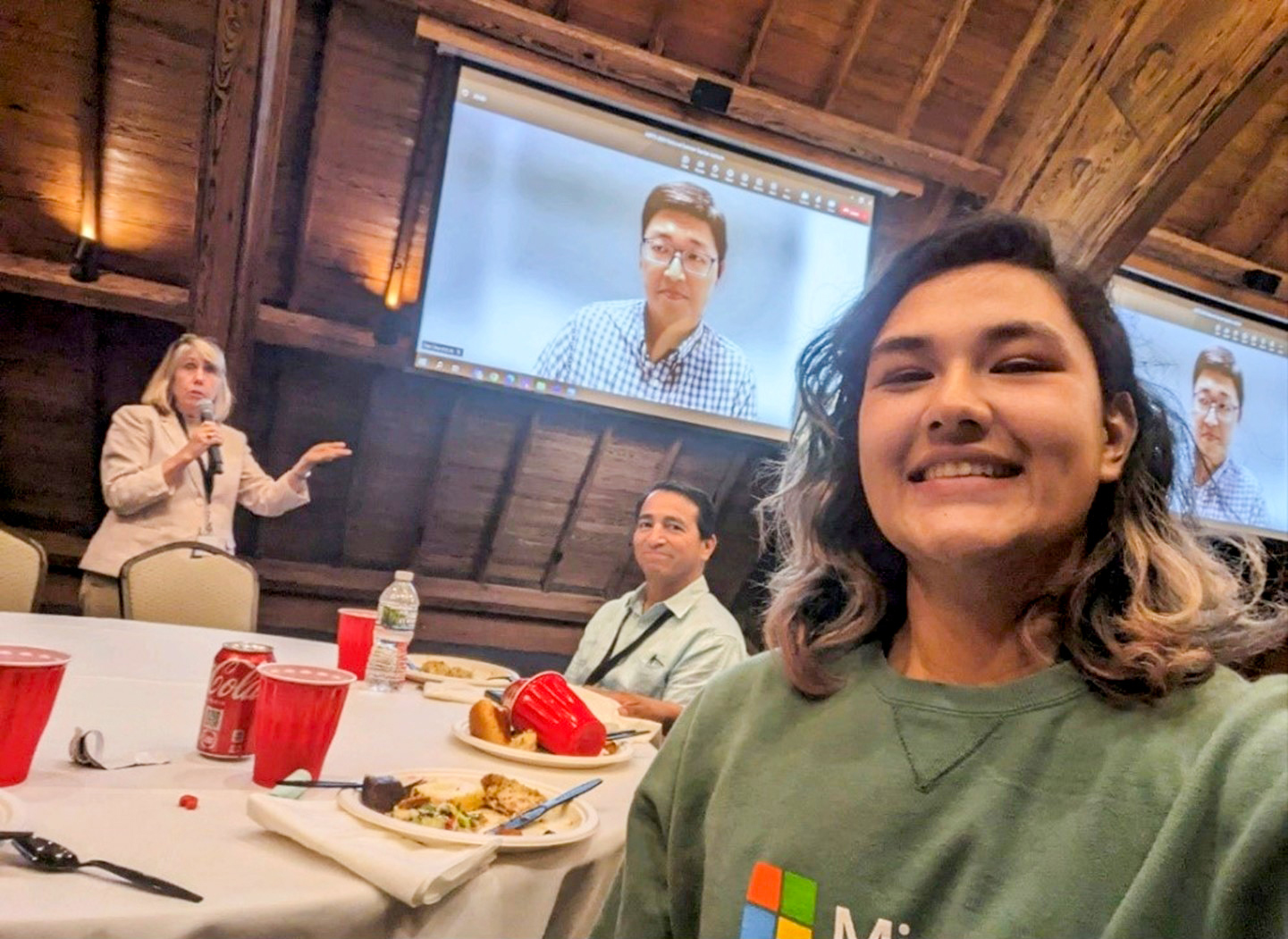 A young woman takes a selfie, with a woman speaking into a microphone in the background, and seated at a dinner table between the two is a smiling man who is her teacher. In the far background, you see a video picture of a man.