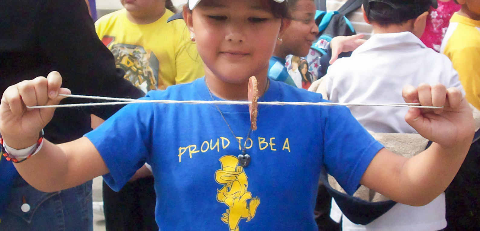 A picture of young Katia Avila Pinedo playing with a string and spinning disk toy and wearing a blue shirt, in with a crowd of elementary school students in the background.