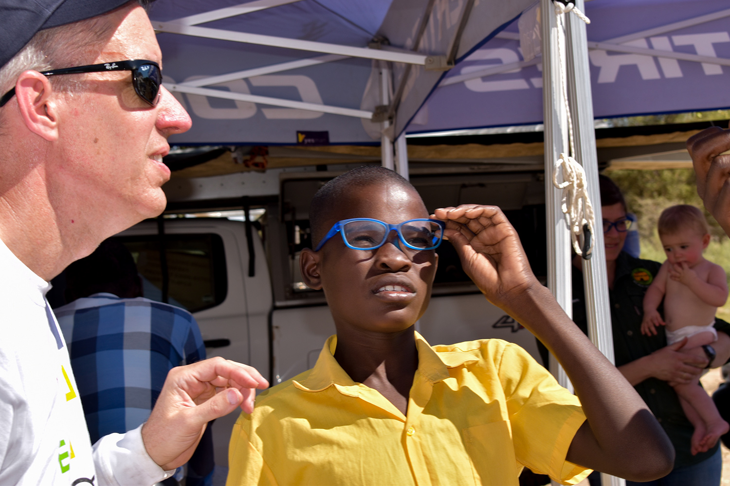 A man in a white shirt helps a young man wearing a yellow with his new prescription blue-framed eyeglasses.