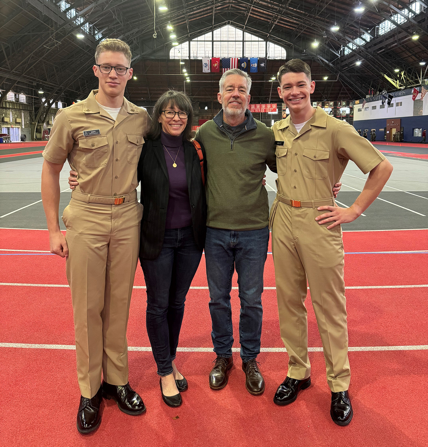  Two young male military recruits in uniform stand next to their parents, a woman with dark hair wearing a black cardigan and a man with white hair wearing a sweater and jeans.