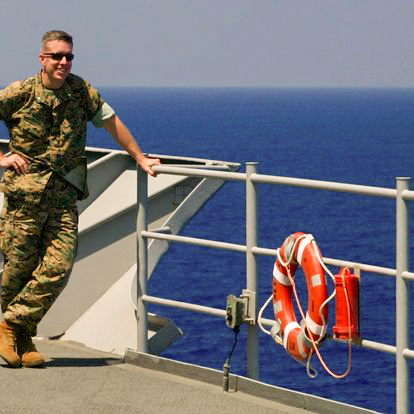 A man in a United States military uniform stands on the deck of a military vessel that is out to sea.