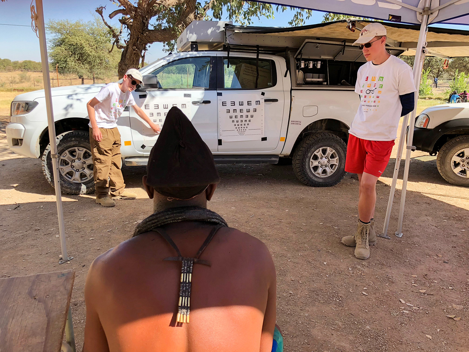 A teenage boy wearing a white shirt gestures to an eye chart affixed to a 4x4 truck and looks at a man sitting in a chair who is having his vision tested. Another teenage boy observes the testing procedure.