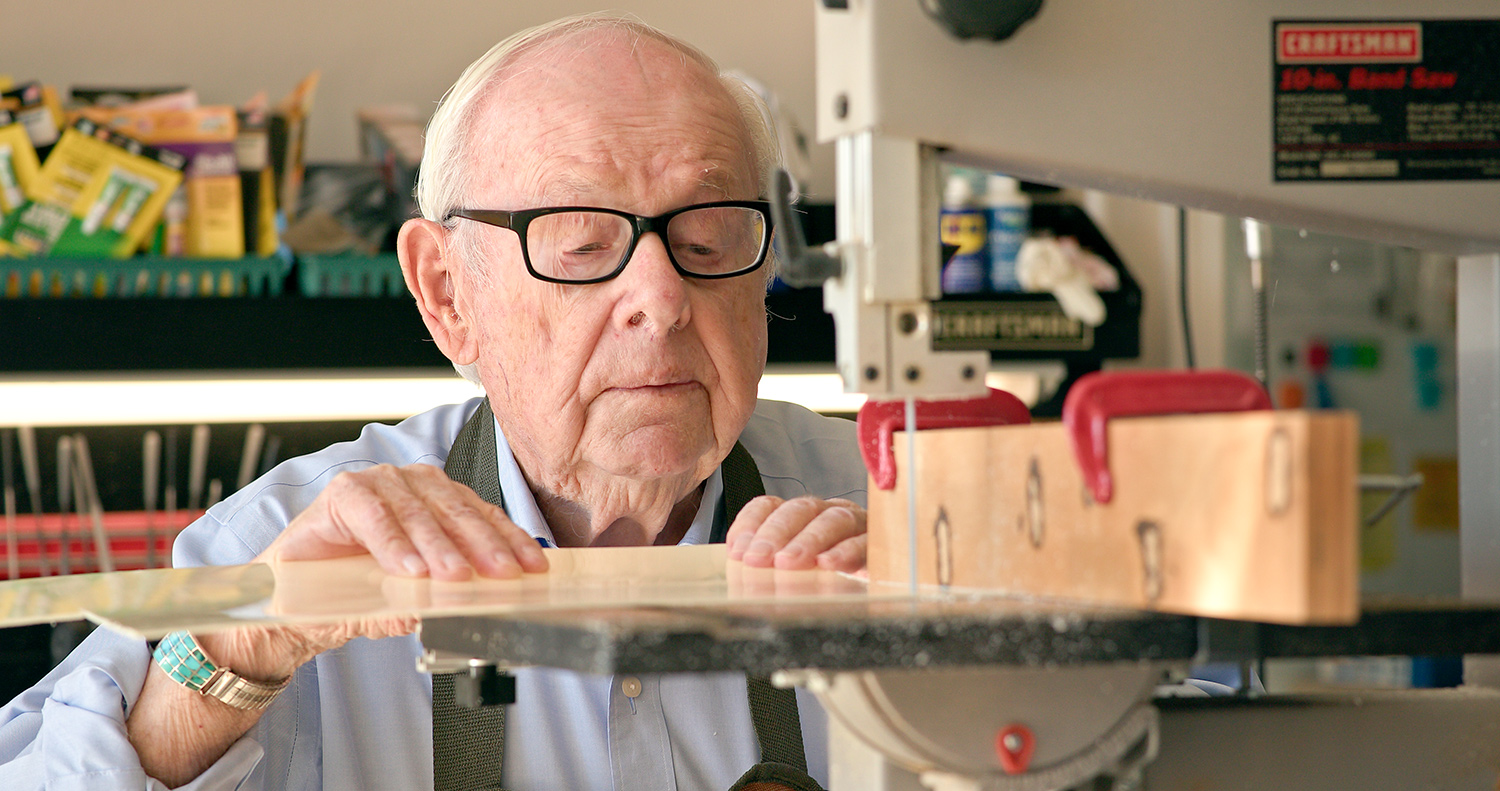 Elderly man with white hair and wearing a blue shirt and work apron operates a bandsaw.