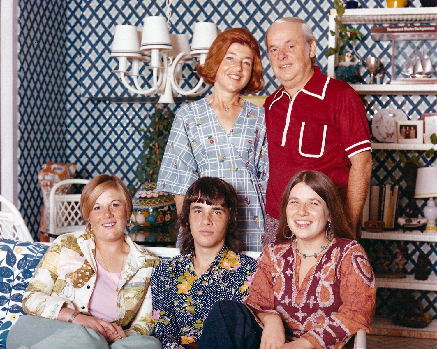 An adult woman wearing a blue dress stands next to a man wearing a red short-sleeve shirt while their three children, a teenage girl, teenage male, and another teenage girl are seated together in front of the parents. 