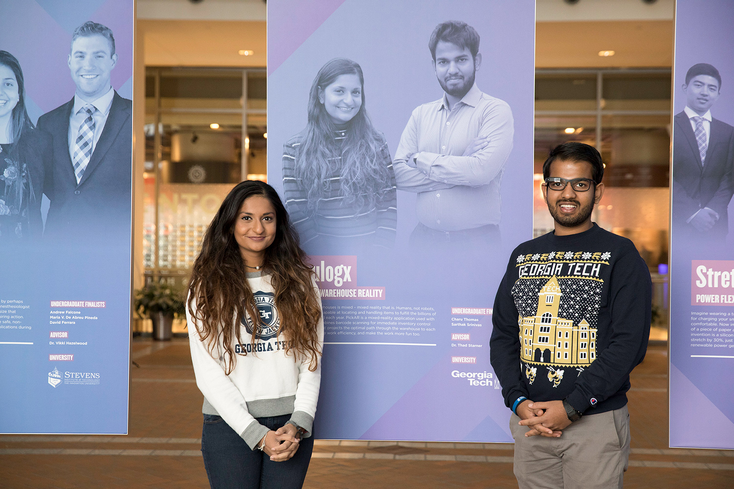 Woman wearing a white sweater stands next to a man wearing a black sweater posing in front of a display with their likeness printed on it.