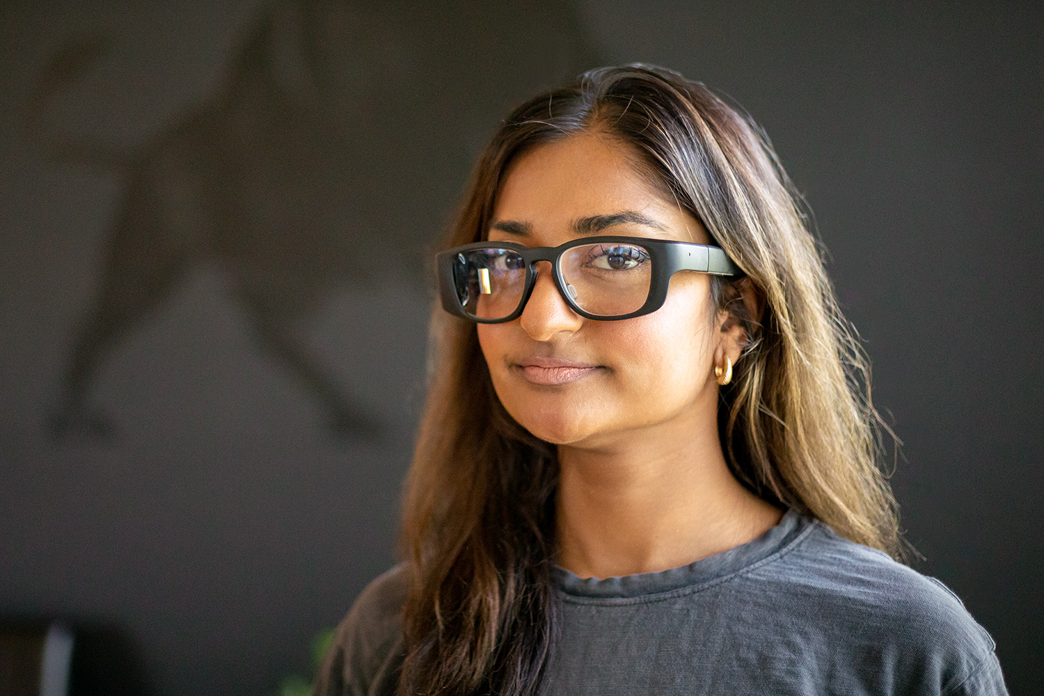 Woman with long dark hair wearing smart glasses poses for photo in front of an image of an ox on the wall.