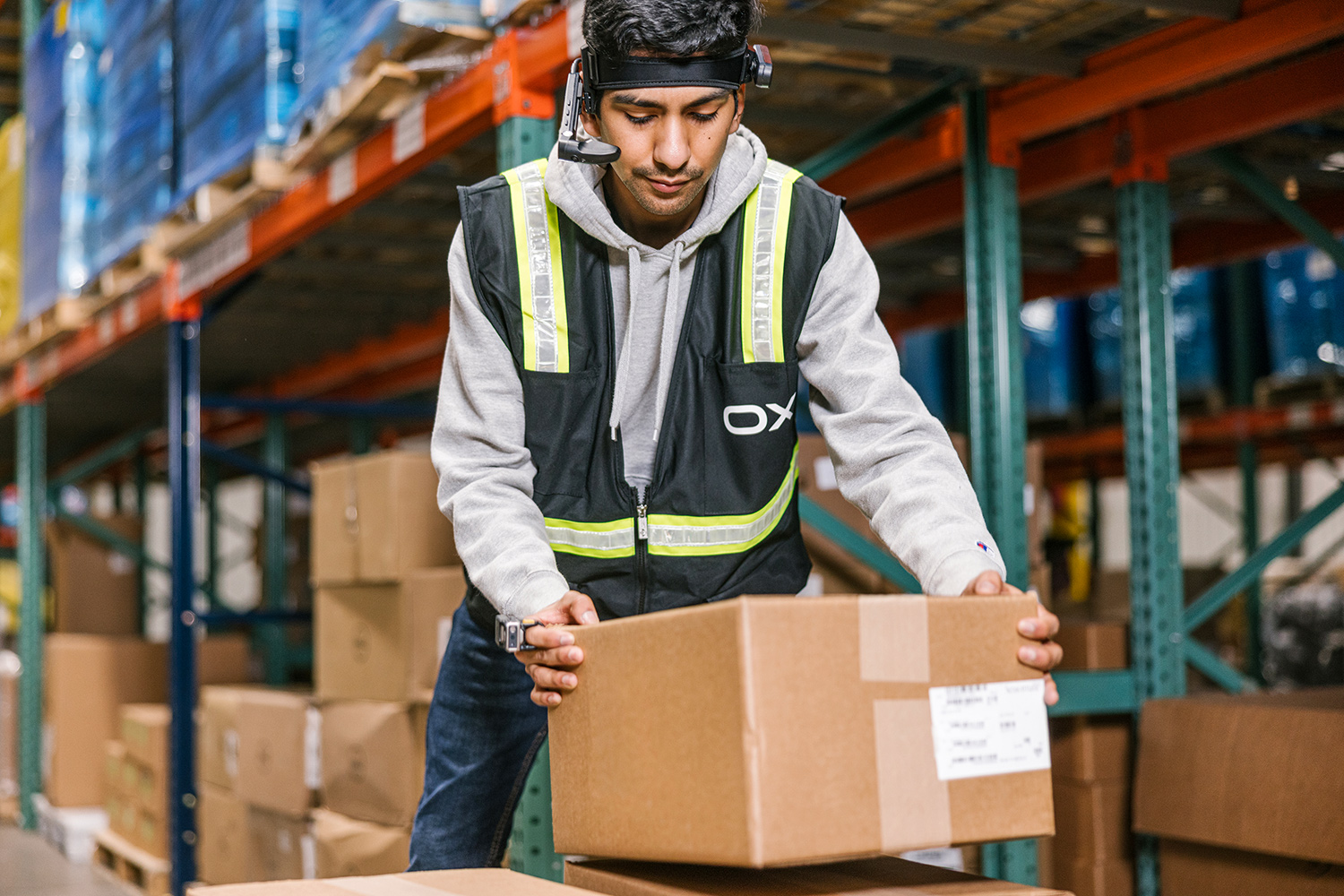 Man wearing a safety reflective vest and smart glasses holds a cardboard box while standing in a warehouse.