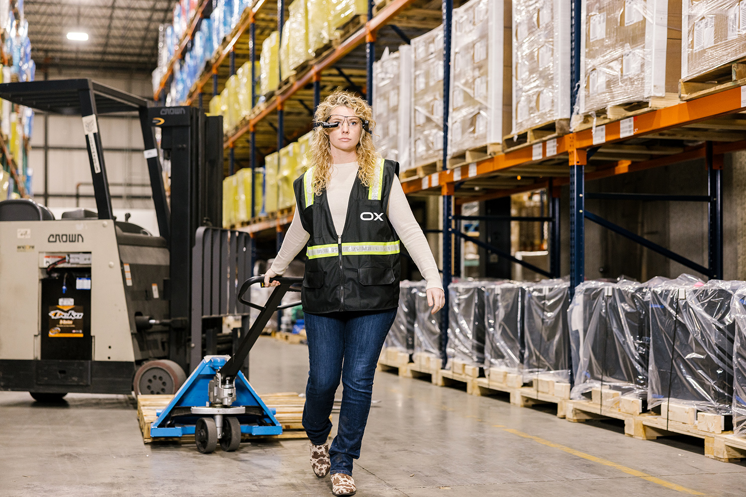 Woman wearing a safety reflective vest pulls a handcart through a warehouse filled with merchandise. 