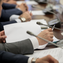 A row of people are seated at a boardroom meeting table. Hands are holding papers. 