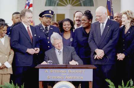 Then-President Bill Clinton signs an appropriations bill that includes the Patent and Trademark Office Efficiency Act into law, in the Rose Garden on November 29, 1999. Clinton is seated and pictured in the middle of the image, surrounded by police and other officials who are standing. The desk Clinton is using to sign the bill says on the front, 'a strong budget for a strong America.' The photo was taken by David Scull and provided courtesy of the William J. Clinton Presidential Library.