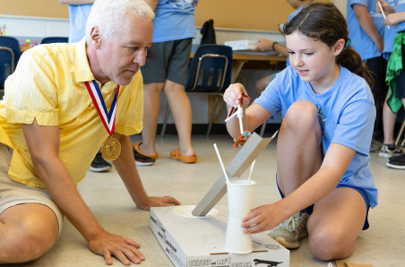 Robert Bryant in a yellow polo shirt and khaki shorts with a National Inventors Hall of Fame medal around his next observes a young girl with brown hair, blue shirt, and blue shorts explain her project in a classroom