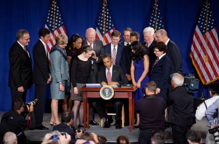 A man in a suit and tie sits at a desk on a stage surrounded by a group of adults and two girls as their photo is taken.