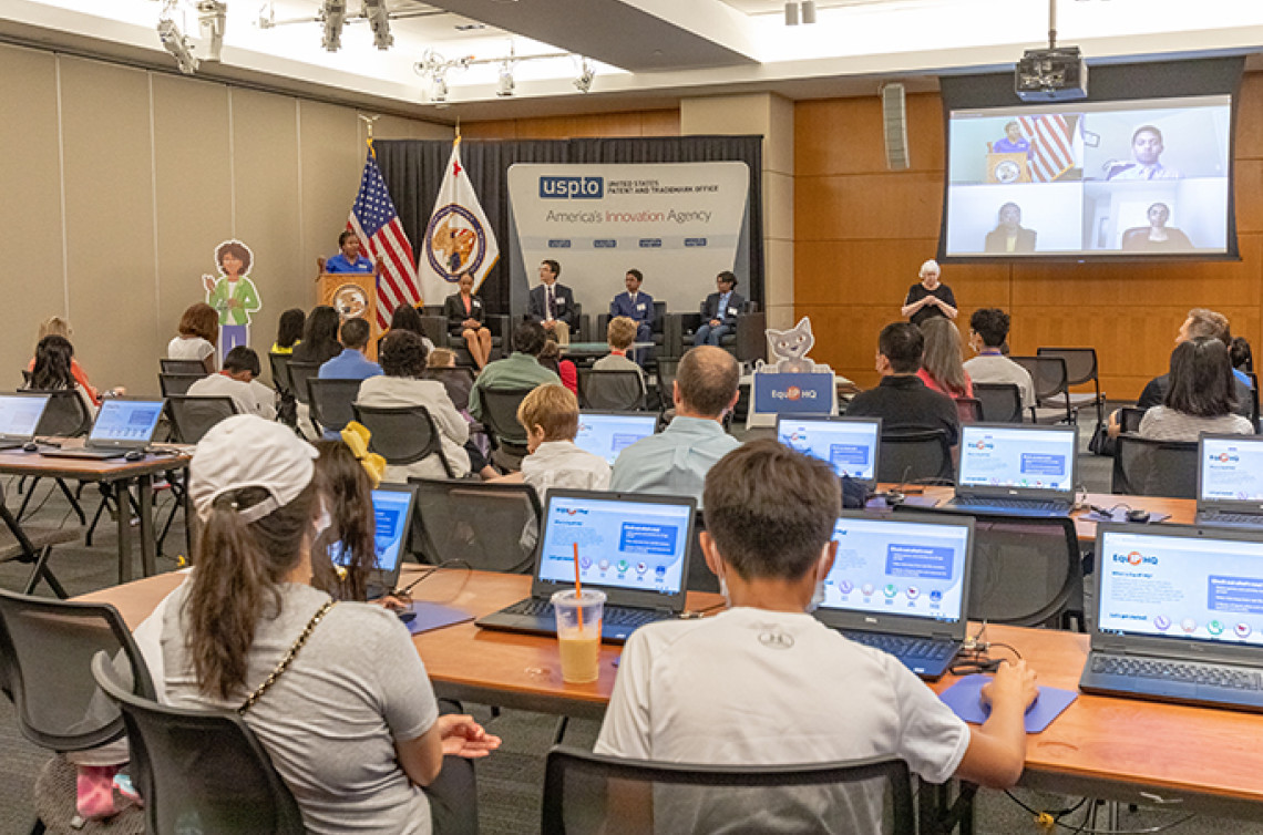 A mix of kids and adults sit in lecture style room with laptops on their desk while a woman speaks at a podium