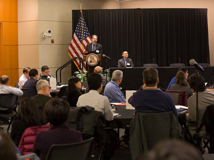 Professionals sit at roundtables while listening to a speaker standing on a stage behind a podium