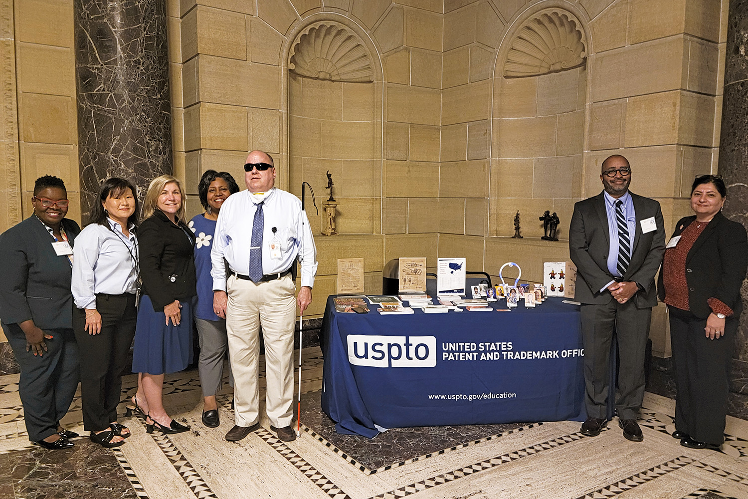 USPTO employees gather around the agency’s table on September 17 at the National Disability Leadership Summit in Washington, D.C., at the Department of Commerce headquarters building. The employees are standing in two groups, on both sides of the table. This photo was taken by Sahar Javanmard from the USPTO.