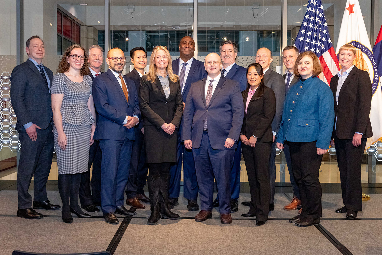 About a dozen people in suits stand for a portrait in front of the U.S. and USPTO flags.