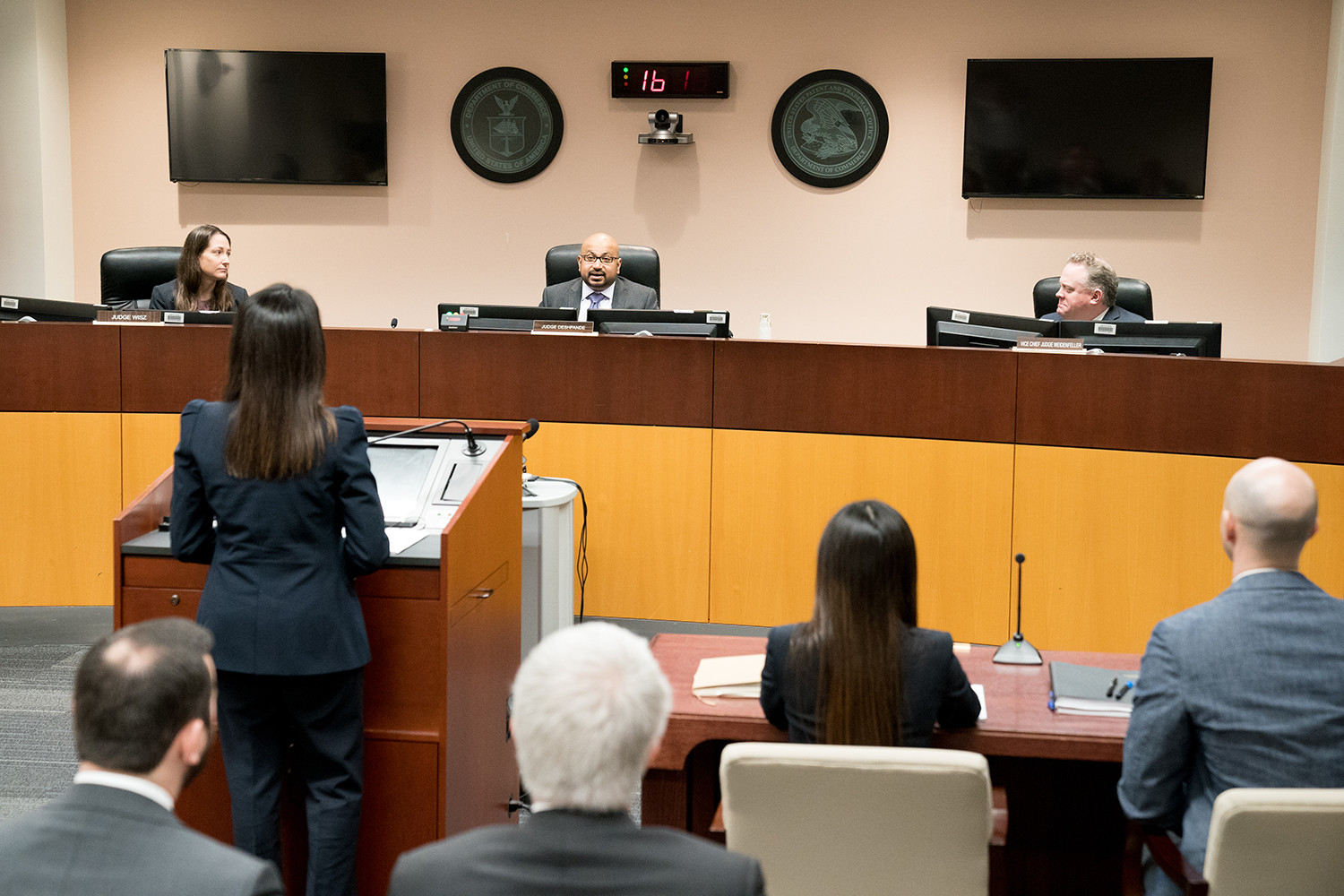 With her back to the camera, a woman appears before three people seated in front of her on a dais. 