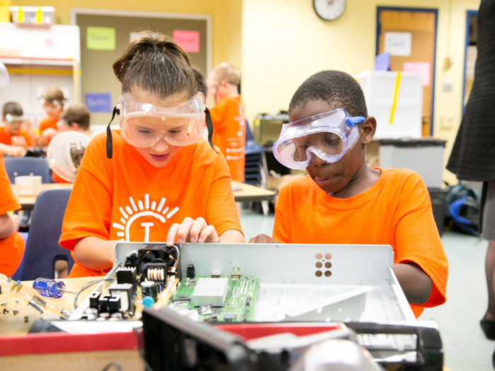 Two Camp Invention students wearing safety googles work on disassembling electronics.
