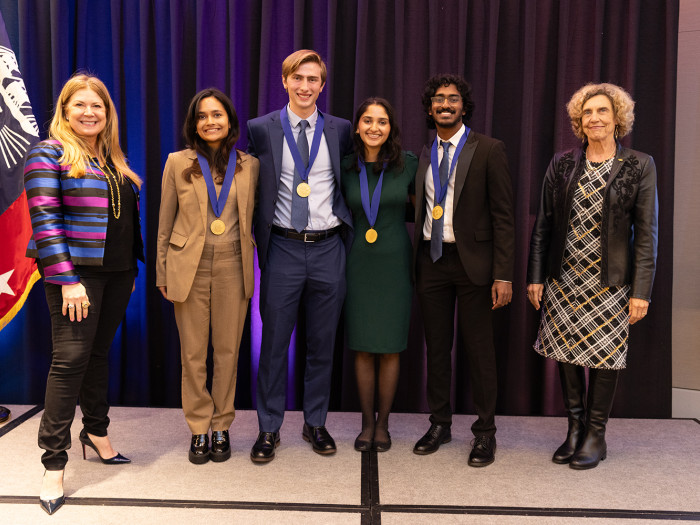 Undergraduate students, two women and two men, stand on stage with USPTO Director Kathi Vidal and Na
