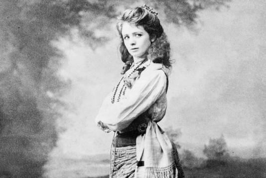 Maude Adams wearing a tunic, skirt, sash, and beads stands at three-quarters profile, framed by trees in the background, and looks off to the right.