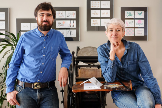 Lex and Santiago pose on either side of a vintage letter press with framed cards in the background. 