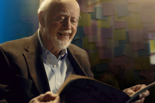 Art Fry, the inventor of post it notes, looks down at a book. He is smiling. In the background, behind Art, post it notes are attached to a wall.