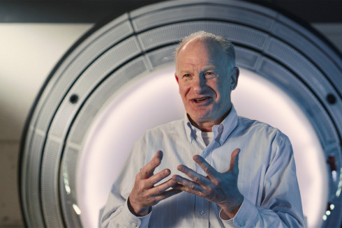 A photo of John Adler in front of medical equipment. Neurosurgeon, inventor, and surfing enthusiast John Adler uses his understanding of energy and desire to help patients to patent a system that can provide therapeutic radiation while protecting healthy tissue.