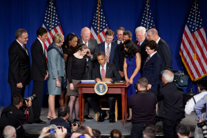 A man in a suit and tie sits at a desk on a stage surrounded by a group of adults and two girls as their photo is taken.