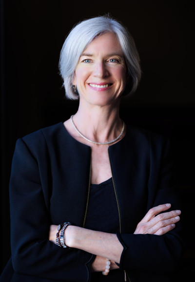 A portrait of a woman with white hair and blue eyes, Jennifer Doudna, smiling with her arms crossed.