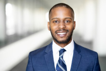 John, a man with black hair and a black beard, in a navy blue suit, navy blue striped tie, and white collared shirt, smiles with the USPTO headquarters hallway behind him.